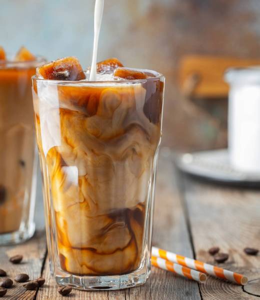 Ice coffee in a tall glass with cream poured over, coffee ice cubes and beans on a old rustic wooden table. Cold summer drink with tubes on a blue rusty background.